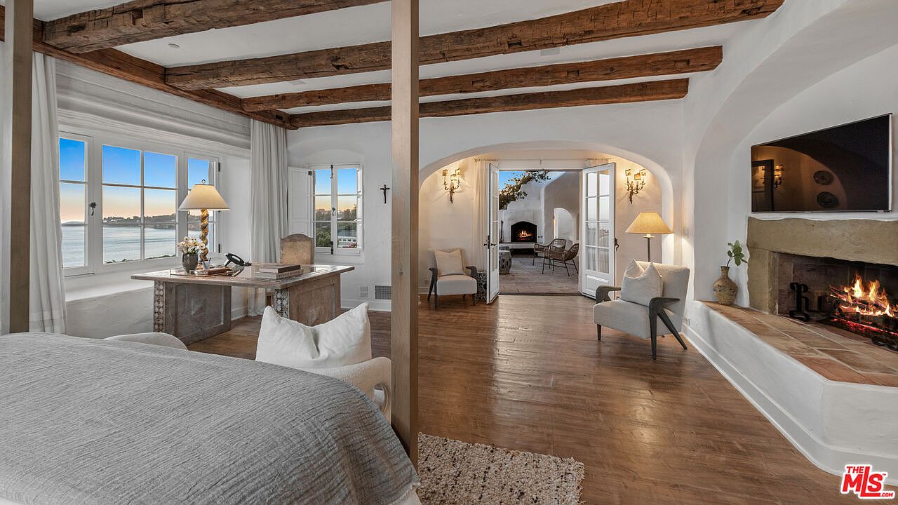 This is a primary bedroom featuring a bed, a desk by the window, and a fireplace with a TV above it. The room has exposed wooden beams on the ceiling and an arched doorway leading to an outdoor patio with a fireplace. The style is rustic and luxurious, creating a warm and inviting atmosphere.