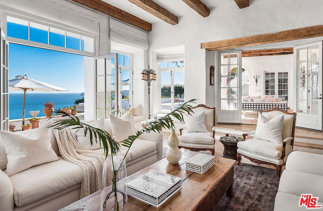 This is an interior shot of a luxurious living room with an ocean view. The room features white sofas and armchairs, wooden ceiling beams, and large windows and doors that open to a balcony overlooking the ocean. The style is elegant and coastal, creating a bright and airy atmosphere.