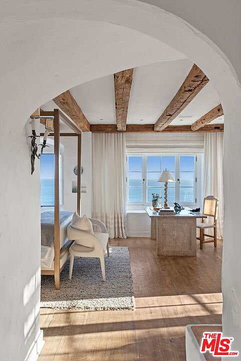 This interior shot showcases a primary bedroom with a view of the ocean. The room features exposed wooden beams, a four-poster bed, and a desk area by the window. The style is coastal and airy, with white walls and curtains creating a bright and inviting space.