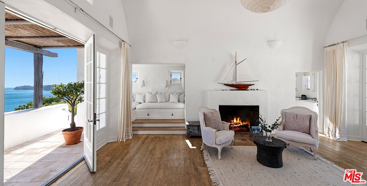 This is an interior shot of a bright and airy living room featuring white walls, hardwood floors, and a fireplace. The room opens onto a balcony with ocean views, enhancing the sense of spaciousness and luxury. The decor is minimalist yet elegant, with comfortable seating and tasteful accents.