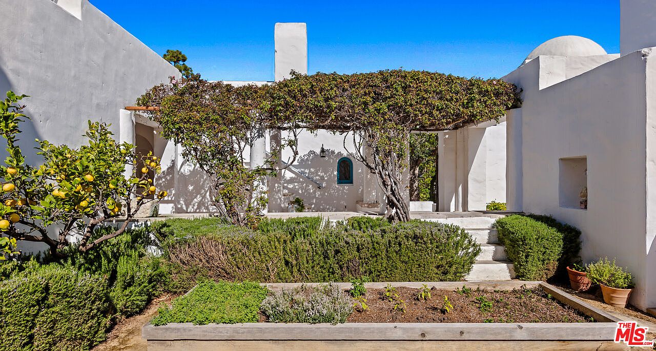 This image showcases a beautifully landscaped yard or garden area, featuring lush greenery, a lemon tree laden with fruit, and a raised garden bed. The backdrop includes a white-walled building with architectural details like a pergola covered in foliage and a small arched window, creating a Mediterranean-inspired aesthetic. The overall impression is serene and inviting, highlighting the property's outdoor living space.