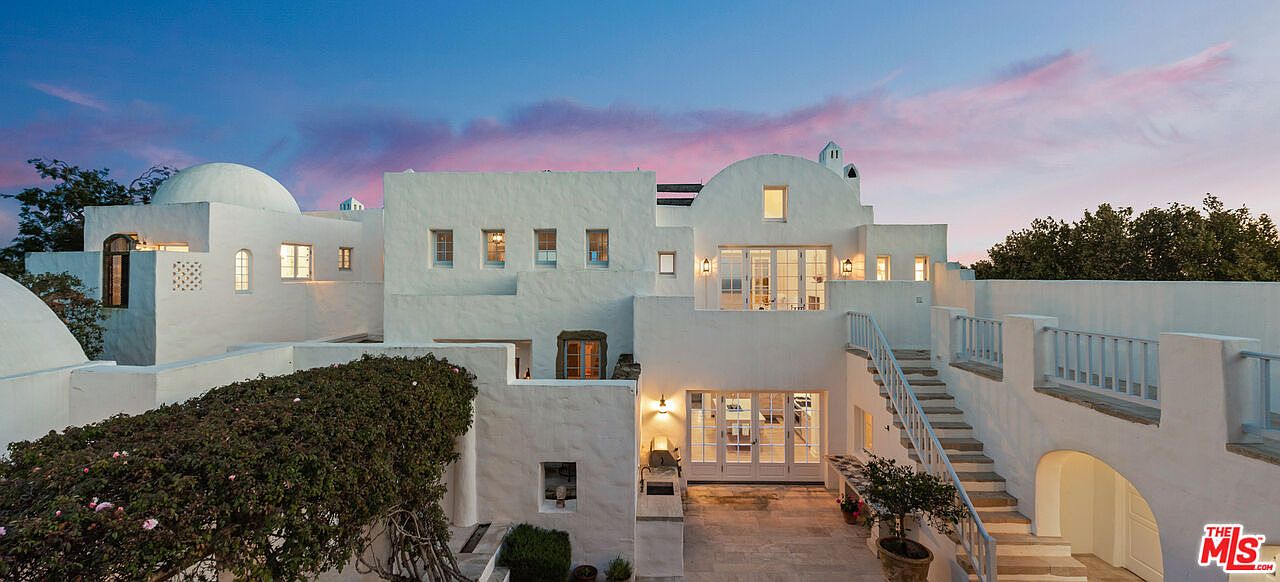 This image showcases the rear exterior of a stunning white stucco home at dusk, featuring a multi-level design with domes, arched doorways, and multiple windows. A staircase leads to an upper balcony, and lush greenery adds to the Mediterranean-inspired aesthetic. The overall impression is one of luxury and architectural uniqueness.