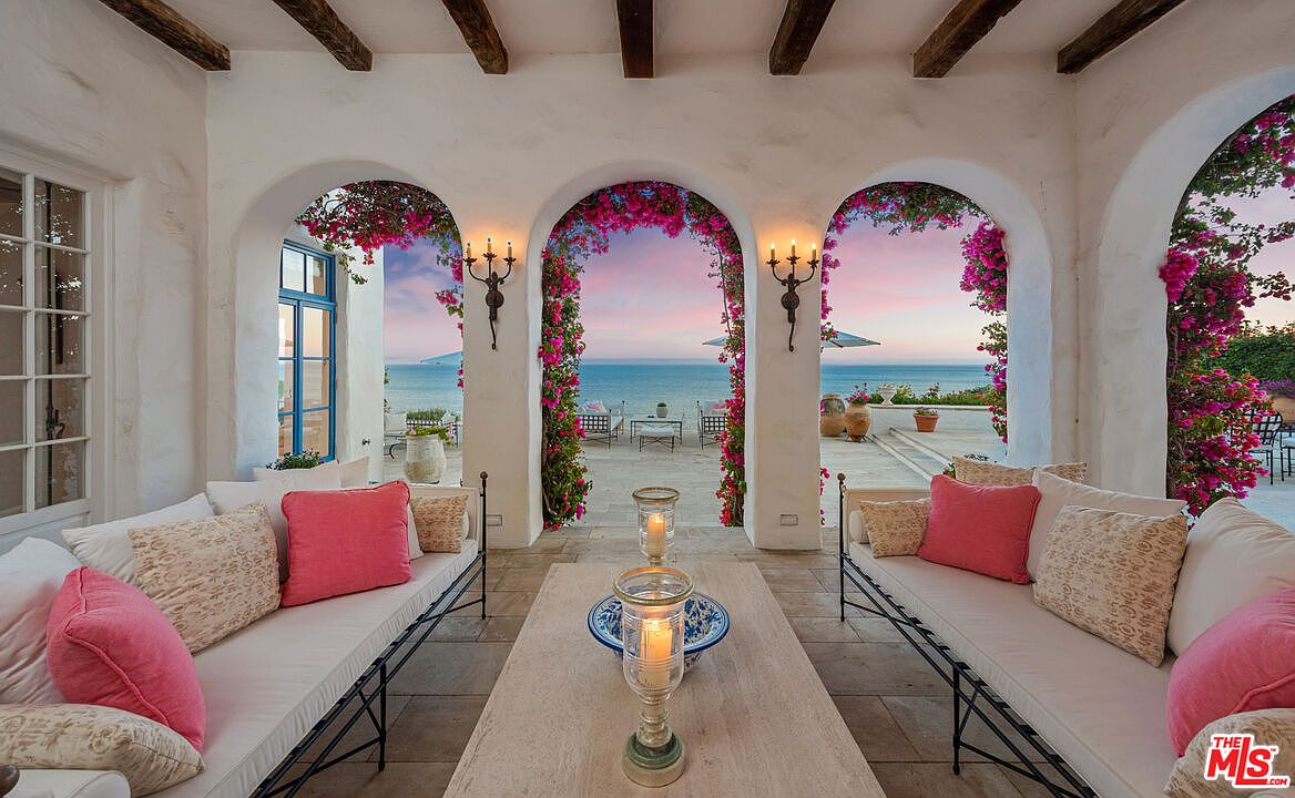 This image showcases a stunning outdoor patio or balcony area with a breathtaking ocean view framed by arched openings adorned with vibrant pink bougainvillea. The space is furnished with comfortable seating arrangements, including cushioned benches and a coffee table, creating an inviting and luxurious atmosphere. The architectural details, such as the exposed wooden beams and sconce lighting, add to the charm and elegance of this outdoor living space.