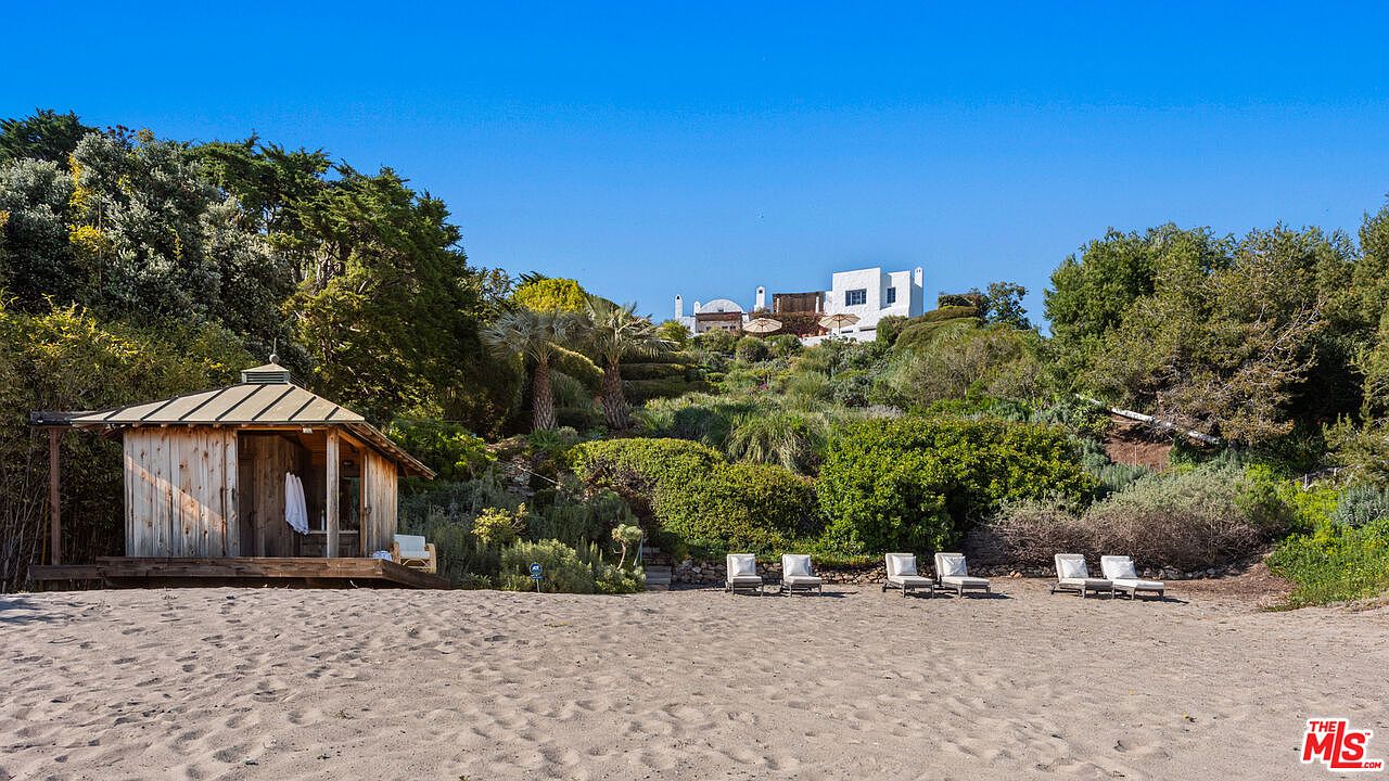 This exterior shot showcases a serene beachfront property with a charming wooden cabana on the sand. Several lounge chairs are arranged on the beach, offering a relaxing space to enjoy the ocean view. The property is landscaped with lush greenery, leading up to a modern white house on a hill, creating a private and luxurious atmosphere.