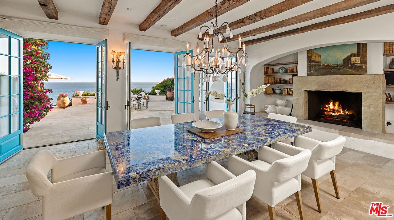 This is an interior shot of a dining room featuring a unique blue marble dining table surrounded by white upholstered chairs. The room opens to an outdoor patio with ocean views through turquoise French doors, and a stone fireplace with a painting above it adds a cozy element. Exposed wooden beams on the ceiling and a decorative chandelier enhance the room's character.