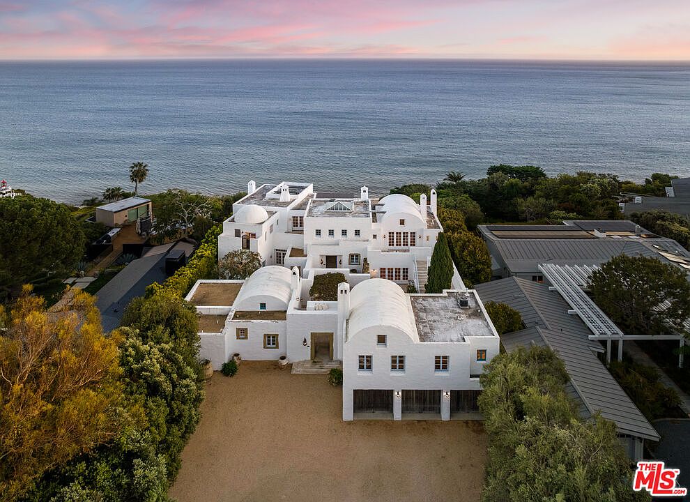 This aerial view showcases a stunning white Mediterranean-style home with arched roofs and multiple levels, set against the backdrop of the ocean. The property features lush landscaping, a spacious front yard, and a clean, modern aesthetic. The overall impression is one of luxury and tranquility, perfect for a high-end real estate listing.