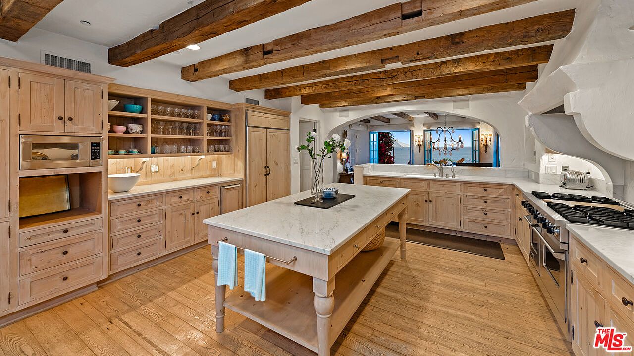 This is a well-lit kitchen featuring wooden cabinetry, a large center island with a marble countertop, and exposed wooden beams on the ceiling. The kitchen includes modern appliances and an open layout that connects to another area with a view of the ocean. The overall style is rustic and elegant.
