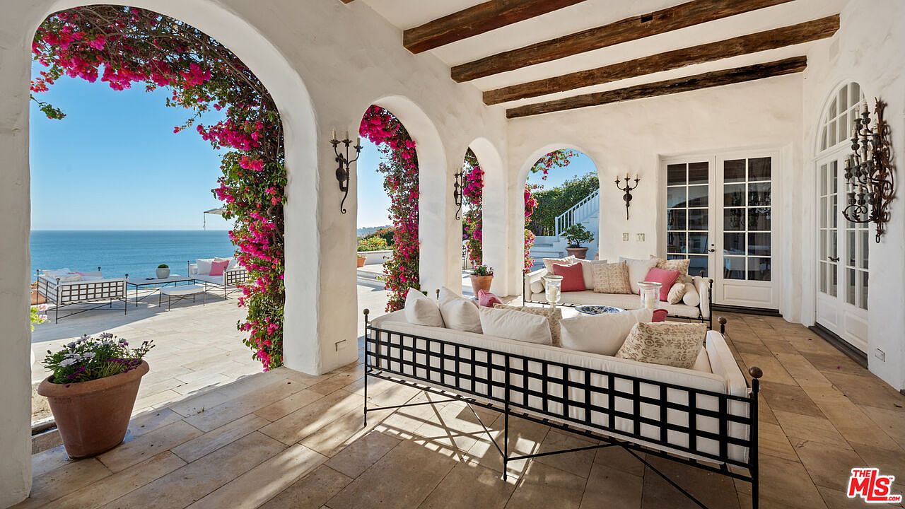This image showcases a luxurious outdoor patio or balcony area with stunning ocean views. The space is defined by elegant white arches adorned with vibrant pink bougainvillea, creating a Mediterranean-inspired ambiance. Comfortable seating arrangements invite relaxation, while the tiled flooring and wooden ceiling beams add to the sophisticated yet inviting atmosphere, perfect for enjoying the coastal scenery.