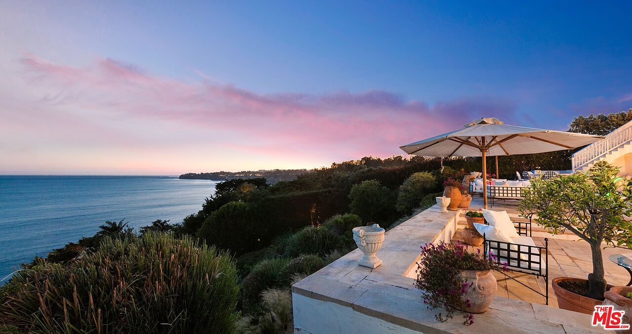 This image showcases a luxurious patio or balcony with stunning ocean views at sunset. The space is furnished with comfortable seating under a large umbrella, potted plants, and decorative urns, creating an inviting outdoor living area. The overall impression is one of tranquility and upscale coastal living.