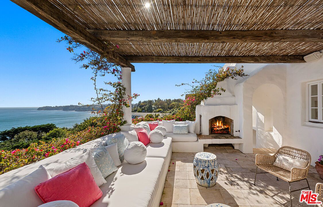 This image showcases a luxurious outdoor patio or balcony area with stunning ocean views. The space features a comfortable white cushioned seating area adorned with decorative pillows, a charming fireplace, and a woven chair, creating an inviting and relaxing atmosphere. A pergola provides shade, while flowering vines add a touch of natural beauty to the scene.