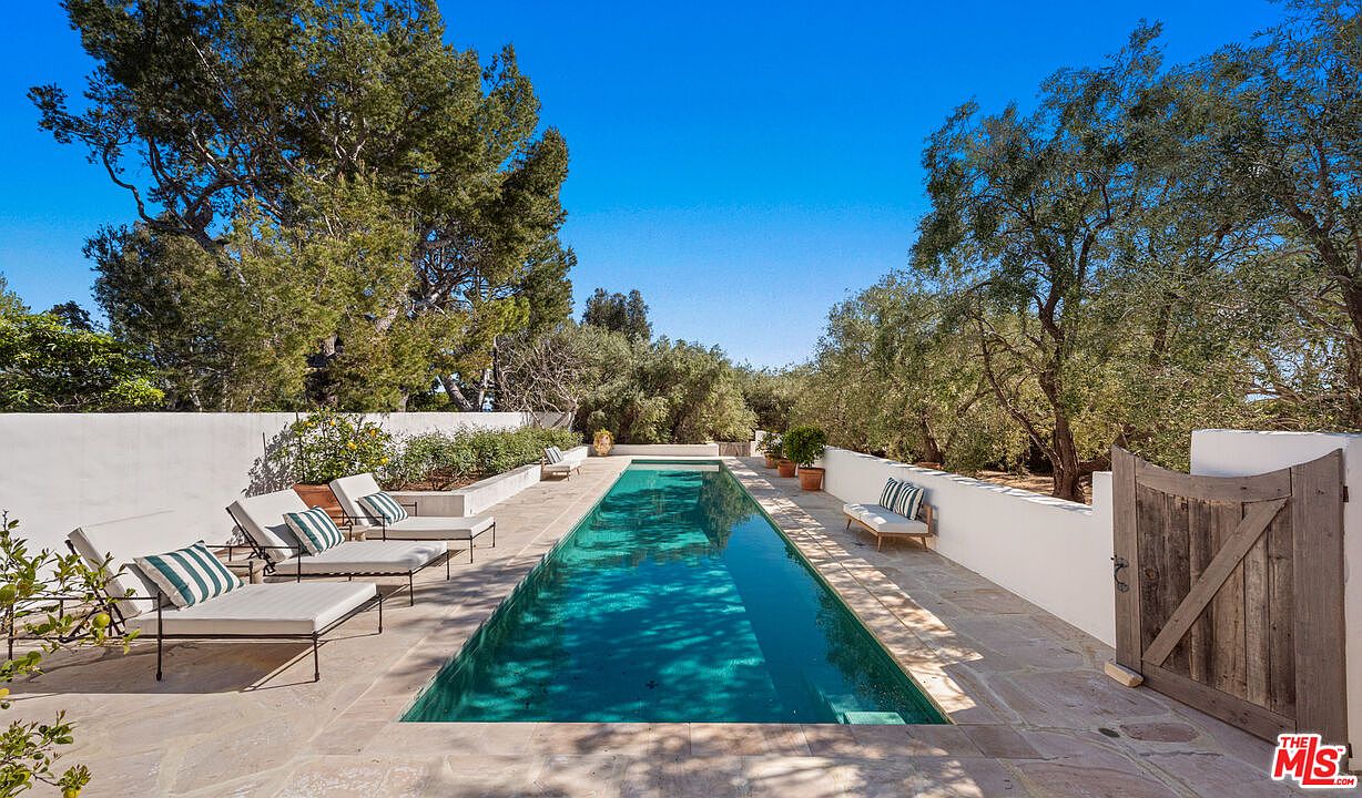 This image showcases a luxurious outdoor pool area, featuring a long, rectangular pool with turquoise water. The pool is surrounded by a stone patio, lounge chairs with striped cushions, and lush greenery, creating a serene and private oasis. A wooden gate and white walls enclose the space, enhancing the sense of seclusion and elegance.