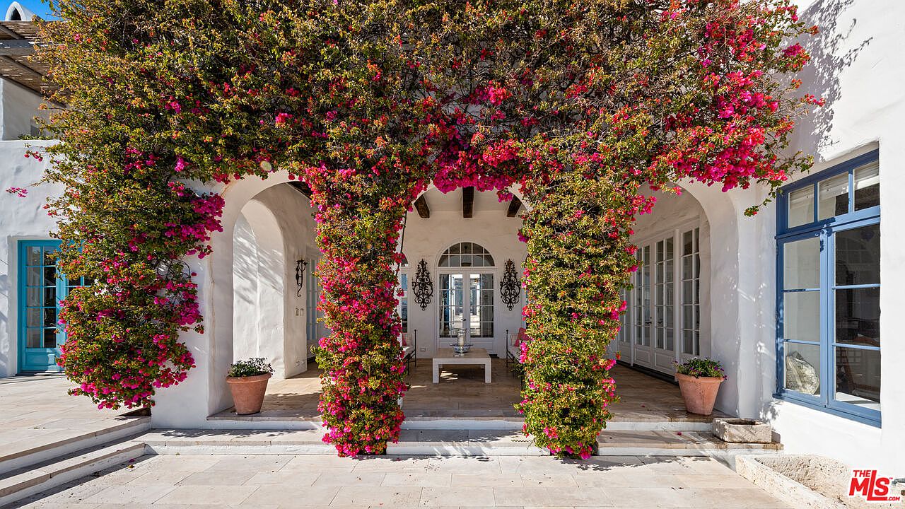 This image showcases the entryway of a white stucco building, beautifully adorned with vibrant pink bougainvillea. The entrance features arched openings and a tiled patio area, creating a welcoming Mediterranean-style ambiance. The blue-framed windows add a pop of color, complementing the overall aesthetic.