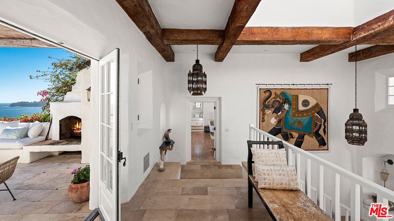 This interior shot showcases a hallway with rustic wooden beams and a view to an outdoor patio with ocean scenery. The hallway features a tiled floor, white walls, and decorative lighting fixtures, creating a blend of Mediterranean and bohemian styles. A tapestry depicting an elephant adds a unique artistic touch to the space.