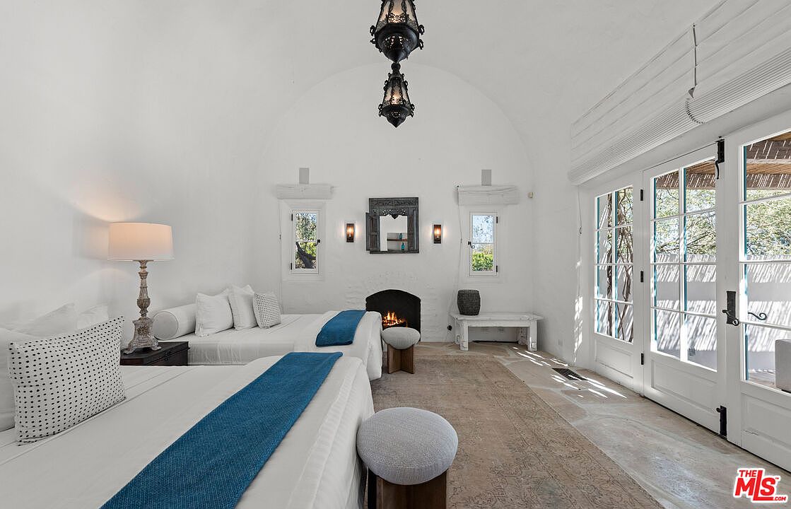 This bedroom features a unique architectural design with a vaulted ceiling and white walls, creating a bright and airy atmosphere. The room is furnished with two beds, each adorned with a blue runner, and complemented by small stools. A fireplace adds a cozy touch, while the French doors provide access to the outside, enhancing the room's appeal.