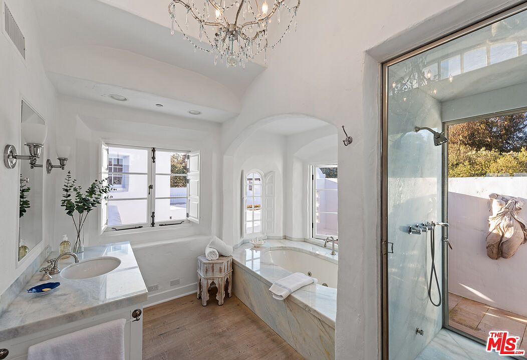 This is a bright and luxurious primary bathroom featuring a standalone tub with marble surround, a glass-enclosed shower, and a vanity with marble countertop. The room is painted in a crisp white, enhancing the natural light from the windows. A chandelier adds a touch of elegance to the space.