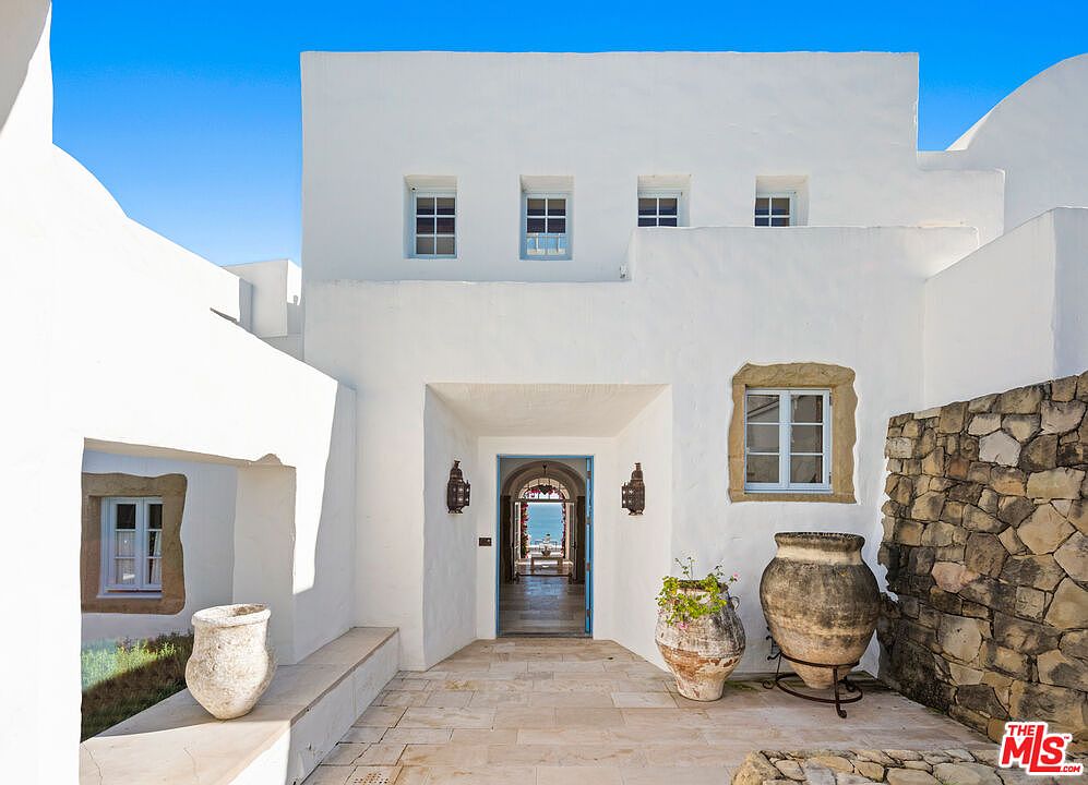 This image showcases the entryway of a stunning white stucco home with a Mediterranean-inspired design. The entrance features a blue-framed doorway leading to a glimpse of the ocean, flanked by decorative lanterns and large terracotta pots. A stone accent wall and tiled patio add to the property's charm and curb appeal.