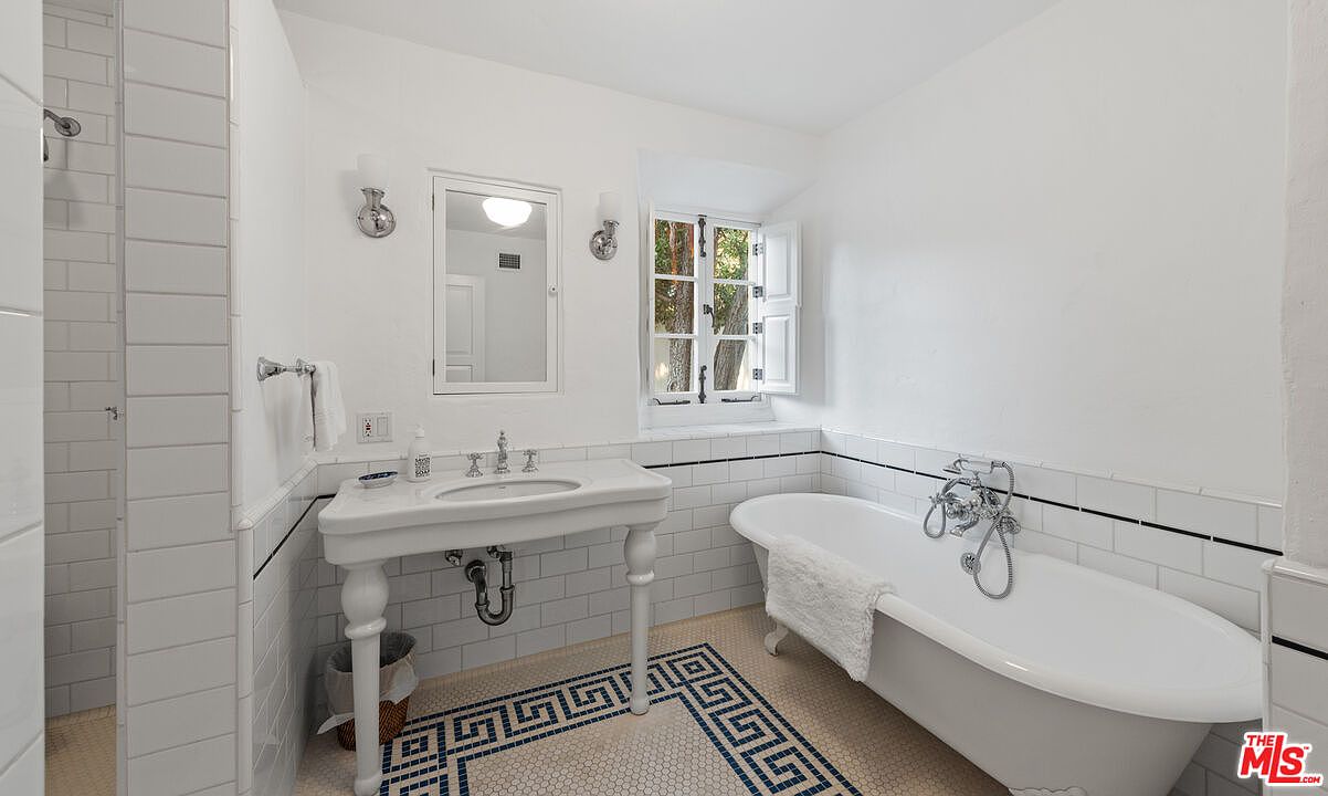 This is a bright and clean bathroom featuring a classic design with white subway tiles, a pedestal sink, and a freestanding bathtub. The floor has a decorative tile pattern, and natural light streams in through a window above the tub. The overall impression is fresh and well-maintained.