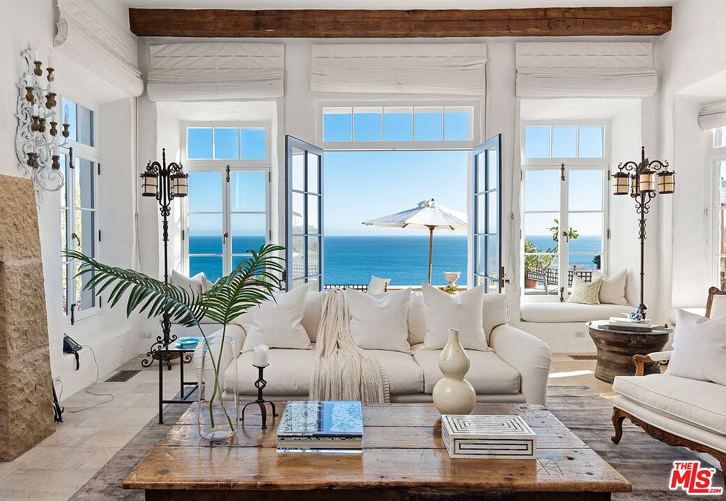 This is an interior shot of a luxurious living room with a stunning ocean view. The room features white walls, large windows, and a comfortable white sofa adorned with pillows and a throw blanket. A rustic wooden coffee table sits in the center, and the overall aesthetic is bright, airy, and coastal chic.
