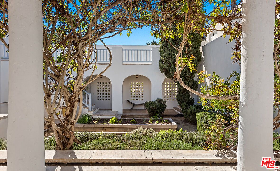 This image showcases a beautifully landscaped yard or garden area, framed by white pillars and lush greenery. The architecture features arched openings with decorative latticework, adding a touch of elegance. The garden is well-maintained with various plants and shrubs, creating a serene and inviting outdoor space.
