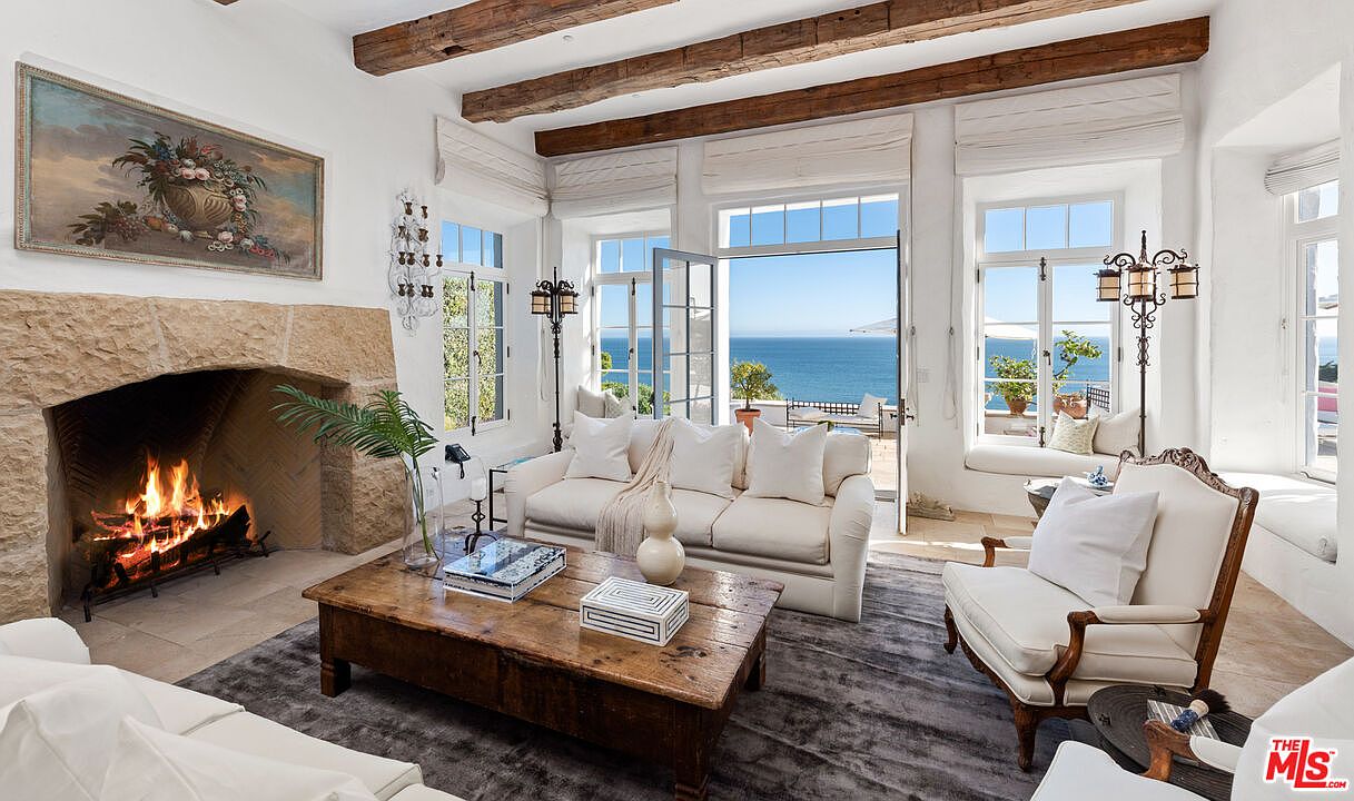 This is an inviting living room featuring a stone fireplace with a roaring fire, a rustic wooden coffee table, and comfortable white sofas and armchairs. Large windows and an open door offer stunning ocean views, enhancing the room's bright and airy atmosphere. Exposed wooden beams add a touch of rustic charm to the otherwise elegant space.