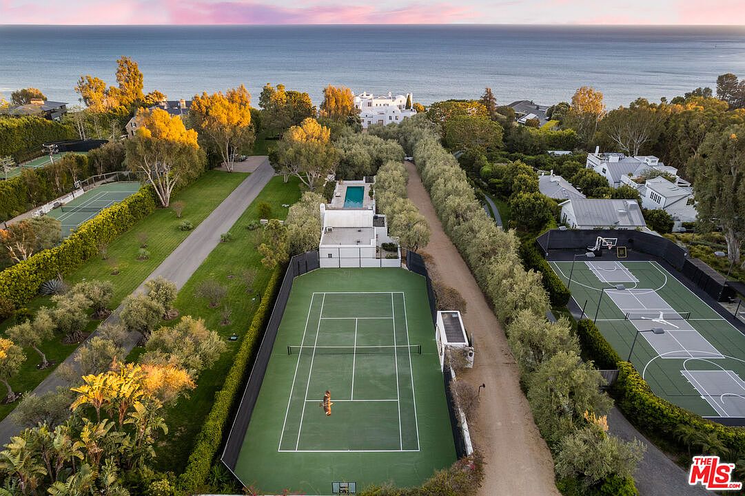 This aerial shot showcases a luxurious estate with multiple amenities, including tennis and basketball courts, surrounded by lush greenery and mature trees. The property features a large main house with a pool and several smaller structures, all set against the backdrop of the ocean. The overall impression is one of privacy, exclusivity, and high-end living.