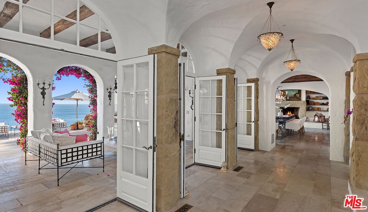 This interior shot showcases a hallway with arched ceilings and stone pillars, leading to an outdoor patio with ocean views. White French doors line the hallway, providing access to the patio. The style is Mediterranean, with a focus on natural light and open spaces, creating a luxurious and inviting atmosphere.