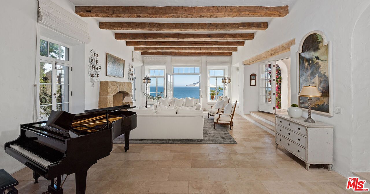 This is a bright and airy living room featuring exposed wooden beams, white walls, and travertine flooring. The room is furnished with a white sofa, armchairs, and a grand piano, creating an elegant and inviting space. Large windows offer stunning ocean views, enhancing the room's appeal.