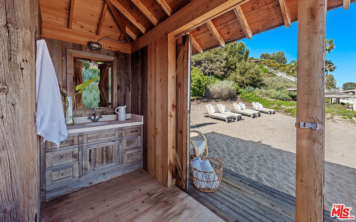 This image showcases a rustic outdoor bathroom or changing area with wooden construction, opening onto a sandy beach. The interior features a vanity with a sink and mirror, while the exterior view includes lounge chairs on the beach and lush greenery in the background. The overall impression is one of relaxed luxury and seamless integration with the natural environment.