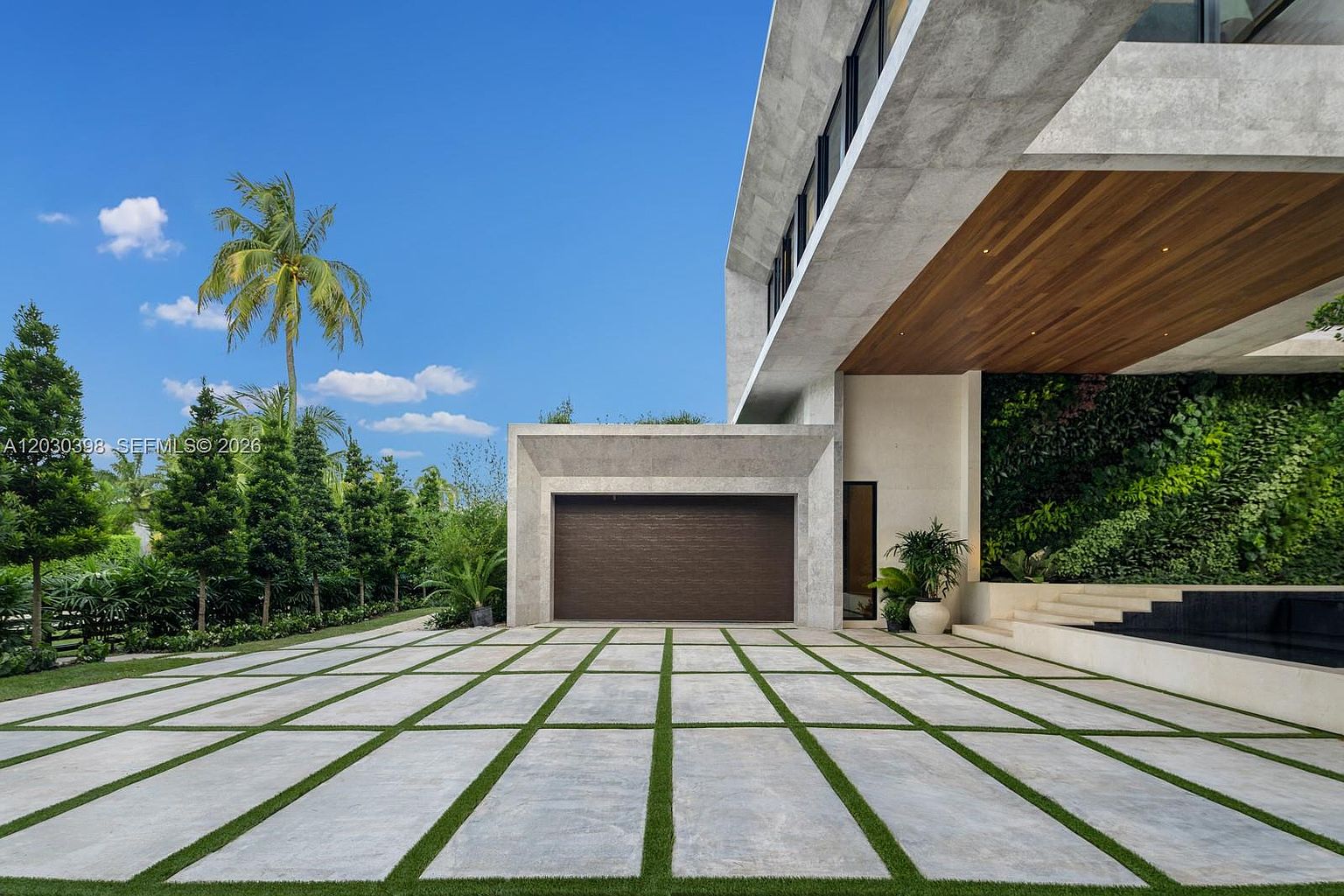 This modern architectural exterior features a prominent garage with a dark, textured door set within a concrete frame. To the right, a lush vertical garden wall adds a vibrant organic element, complemented by a wooden soffit overhead and a tiered stone water feature. The foreground consists of a geometric driveway paved with large concrete pavers separated by strips of green grass, creating a clean, contemporary aesthetic.