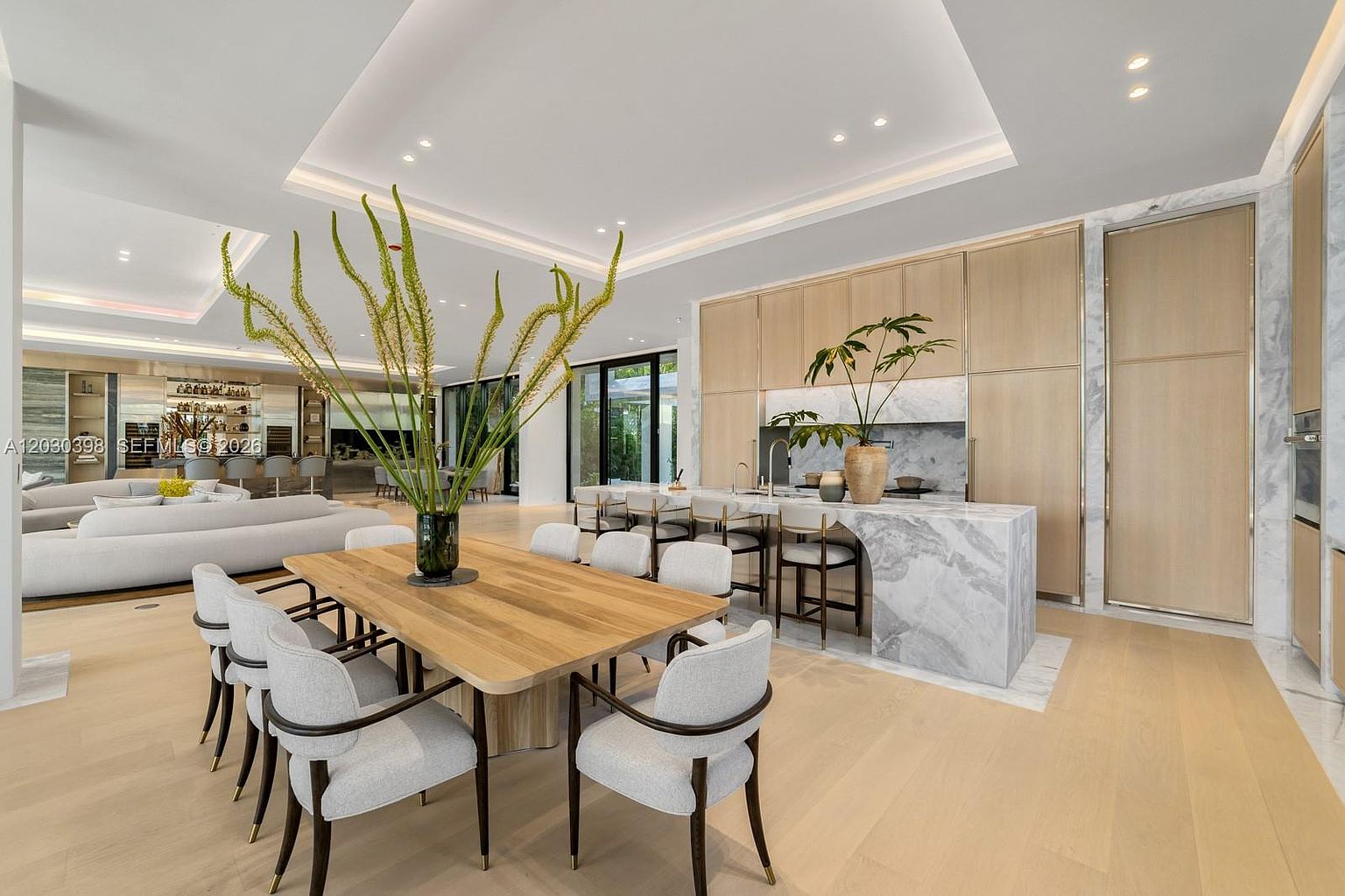 This open-concept space seamlessly blends a dining area with a sophisticated kitchen, featuring light wood cabinetry and a large stone-topped island with integrated seating. The room is illuminated by recessed lighting and large windows that provide a bright, airy atmosphere. The perspective captures the flow between the dining table and the kitchen, highlighting the modern, minimalist aesthetic and high-end finishes throughout.