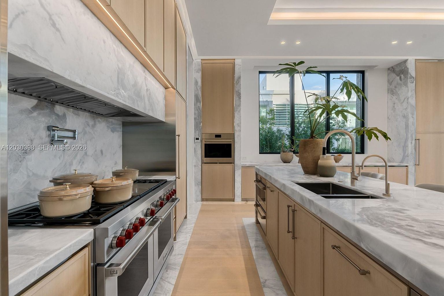 This modern kitchen features a sleek, minimalist aesthetic with light wood cabinetry and expansive stone countertops. A large central island includes a double-basin sink, while the perimeter showcases a professional-grade range with a matching stone backsplash. The space is brightened by natural light from a large window, creating an airy and sophisticated atmosphere.