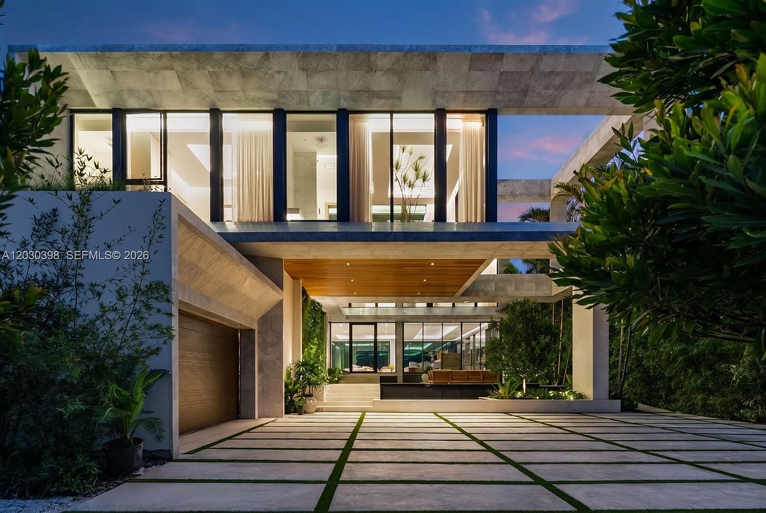This striking modern residence features a bold architectural design with clean lines, a flat roofline, and a mix of stone and wood accents. The front facade showcases large windows that span the upper level, while a covered entryway leads to a recessed front door. A paved driveway with integrated grass strips leads up to the home, framed by lush landscaping and ambient evening lighting that highlights the structure's geometric form.
