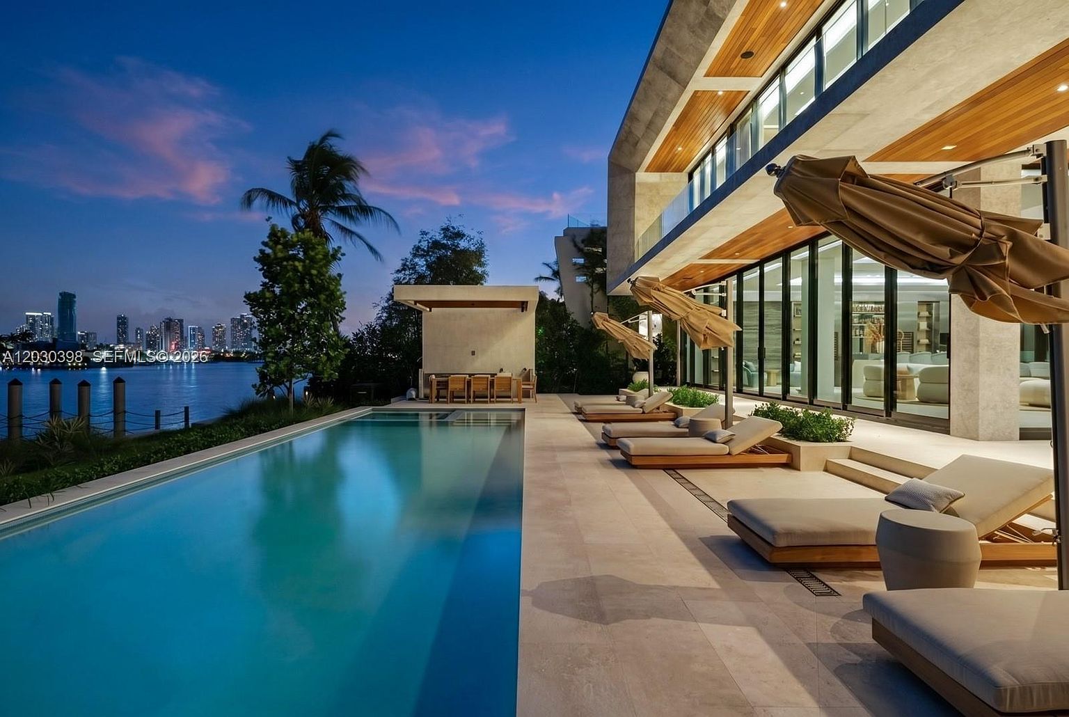 This luxurious waterfront property features a sleek, rectangular infinity-edge pool that stretches toward a scenic bay view at twilight. The expansive stone patio is furnished with multiple lounge chairs and umbrellas, while a covered outdoor dining area sits near the water's edge. The modern architecture is highlighted by warm wood-paneled ceilings and large glass walls that seamlessly connect the indoor living space to the outdoor terrace.