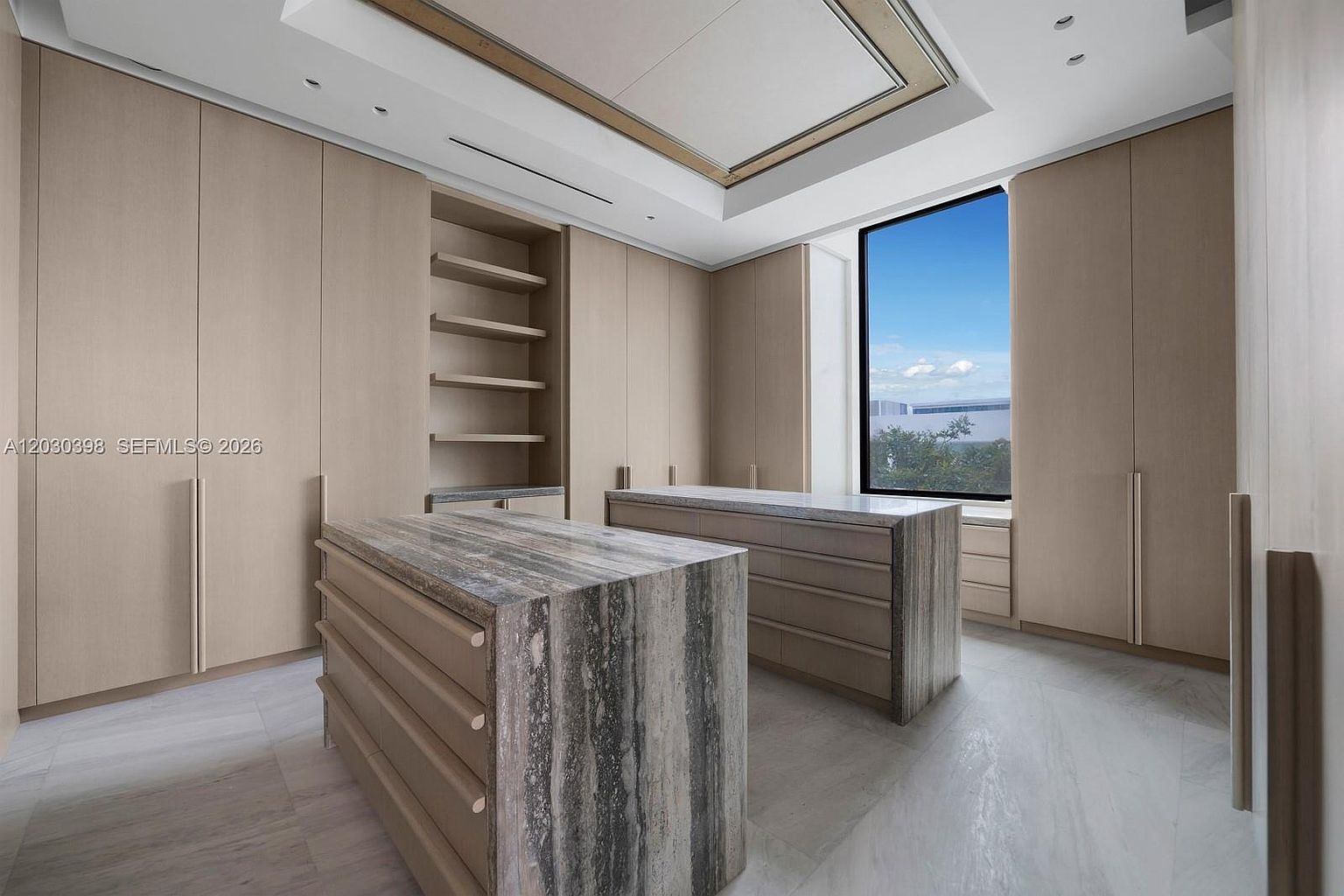This sophisticated walk-in closet features extensive light wood cabinetry and two central islands topped with striking, heavily veined stone. A large window provides natural light and an expansive view, while the neutral color palette and clean lines create a luxurious, organized atmosphere. The perspective is centered, showcasing the symmetrical layout and high-end finishes of this custom storage space.