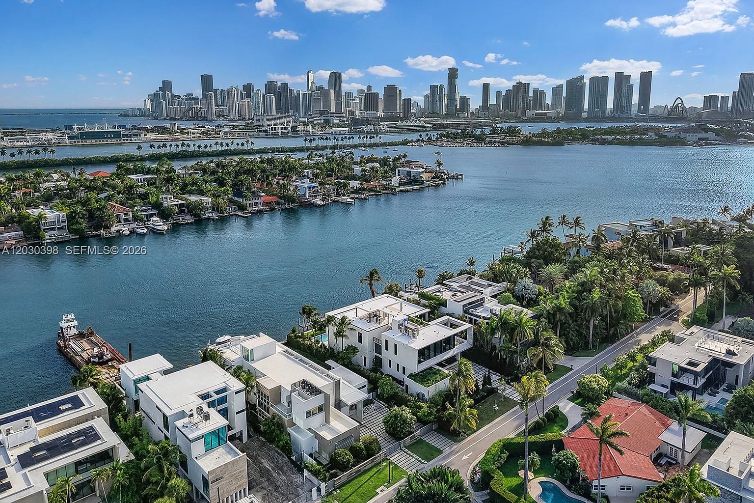 This high-angle aerial view captures a luxurious waterfront neighborhood featuring modern, multi-level residences with flat roofs and expansive terraces. The scene overlooks a calm bay with a vibrant city skyline visible in the distance under a bright, partly cloudy sky. Lush tropical landscaping and private docks line the water's edge, emphasizing the exclusive and serene coastal lifestyle.