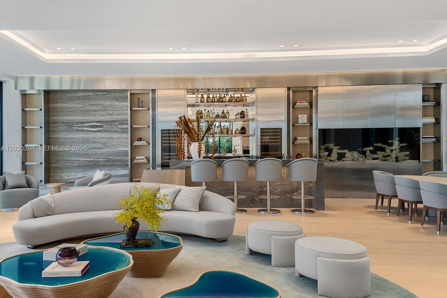 This expansive, open-concept living area features a sophisticated contemporary design with a curved sofa and unique, organic-shaped coffee tables. In the background, a sleek bar area with metallic cabinetry and bar seating serves as a focal point, complemented by a large built-in aquarium. The space is finished with light-toned flooring and recessed lighting, creating a bright and luxurious atmosphere.