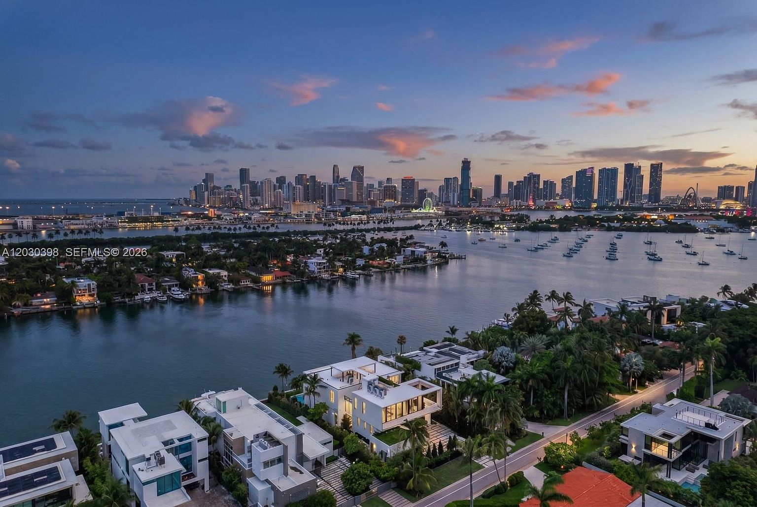 This high-angle aerial view captures a luxurious waterfront residential neighborhood in Miami, featuring modern, multi-level homes with flat roofs and clean architectural lines. The foreground showcases these contemporary estates nestled among lush tropical landscaping, while the background offers a sweeping, panoramic vista of the Miami skyline across the bay at dusk. The scene is illuminated by the soft, warm glow of the setting sun, highlighting the tranquil water and the vibrant city lights beginning to emerge.