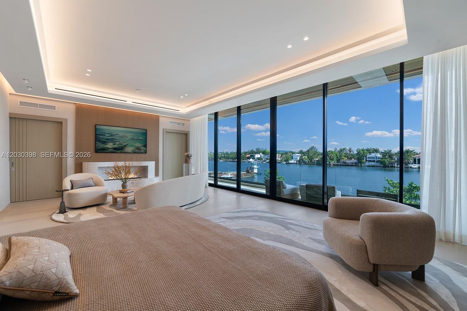 This luxurious primary bedroom features a neutral color palette and expansive floor-to-ceiling windows that provide a panoramic view of the waterfront. The room is furnished with a plush bed, a curved sofa, and a comfortable armchair, all arranged around a sleek fireplace with a minimalist surround. The space is bathed in natural light, creating an airy and sophisticated atmosphere that emphasizes the seamless connection between the interior living space and the outdoor scenery.