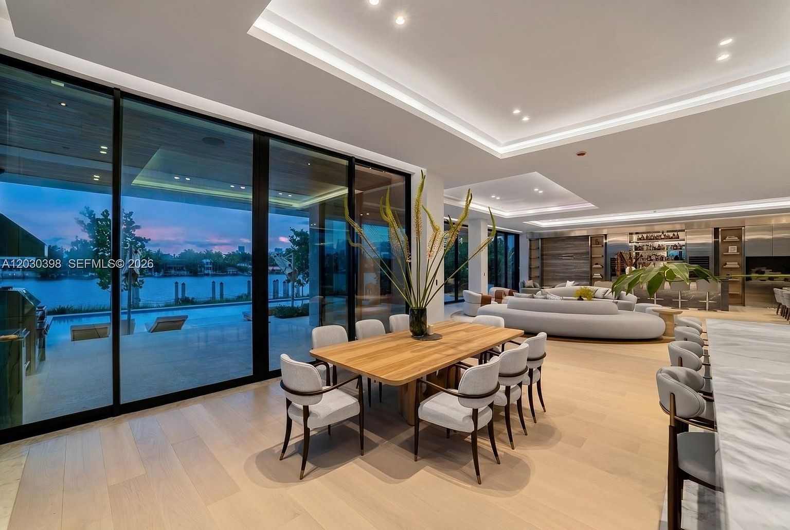 This expansive, open-concept dining area features a large light wood table surrounded by eight upholstered chairs, centered beneath a recessed ceiling with integrated lighting. The space is defined by floor-to-ceiling glass walls that provide a seamless transition to the outdoor pool and waterfront view. In the background, a sophisticated living area with a curved sofa and a sleek bar setup completes the modern, high-end aesthetic of this luxury residence.