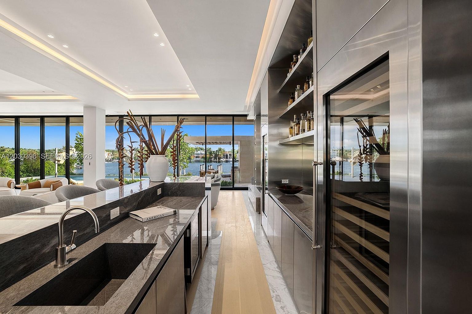 This sleek, modern kitchen area features a long island with a dark stone countertop and an integrated sink, complemented by a dedicated bar section with open shelving and a built-in wine refrigerator. The space is defined by a sophisticated mix of dark stone surfaces and light wood flooring, creating a high-end, contemporary aesthetic. Large, expansive windows provide a seamless connection to the outdoor waterfront view, bathing the interior in natural light.
