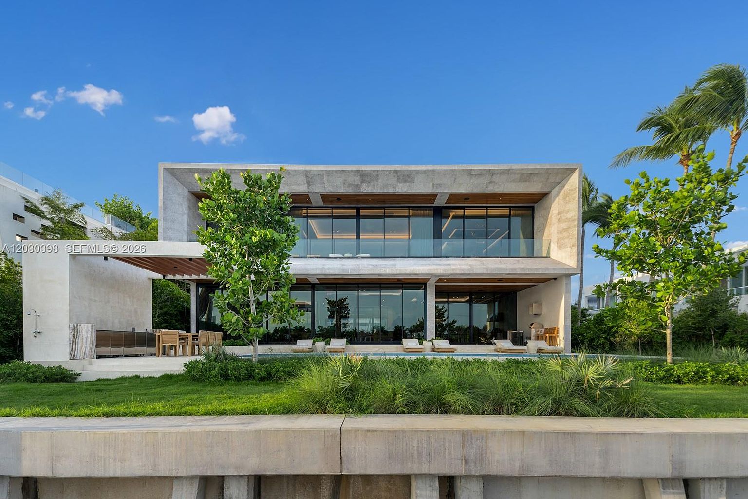 This modern, two-story residence features a striking architectural design with clean lines and a prominent concrete frame. The rear facade showcases extensive floor-to-ceiling glass walls that open onto a covered patio area, complete with an outdoor kitchen and dining space overlooking a swimming pool. Lush landscaping, including mature trees and manicured grass, frames the property, creating a serene and luxurious waterfront atmosphere.