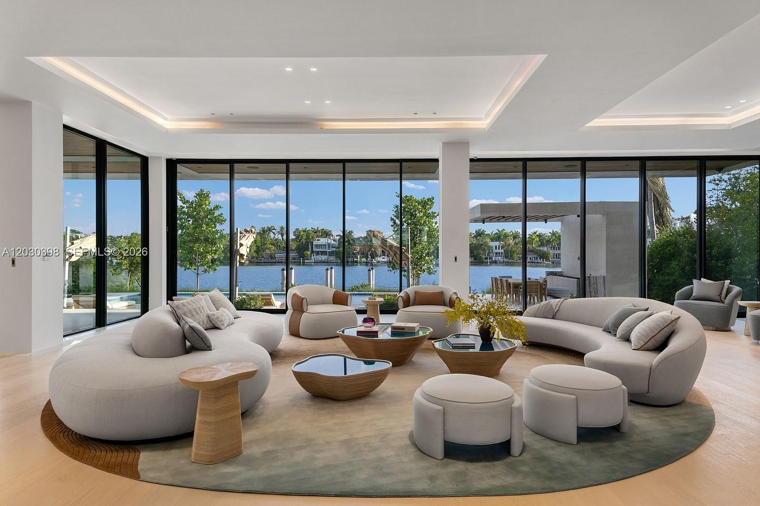 This expansive, modern living room features a curved, neutral-toned sofa arrangement centered around a large, circular area rug. The space is defined by floor-to-ceiling windows that provide an unobstructed view of the waterfront and lush greenery outside. The minimalist aesthetic is complemented by light wood flooring, sculptural coffee tables, and soft, organic furniture shapes that create a serene and luxurious atmosphere.