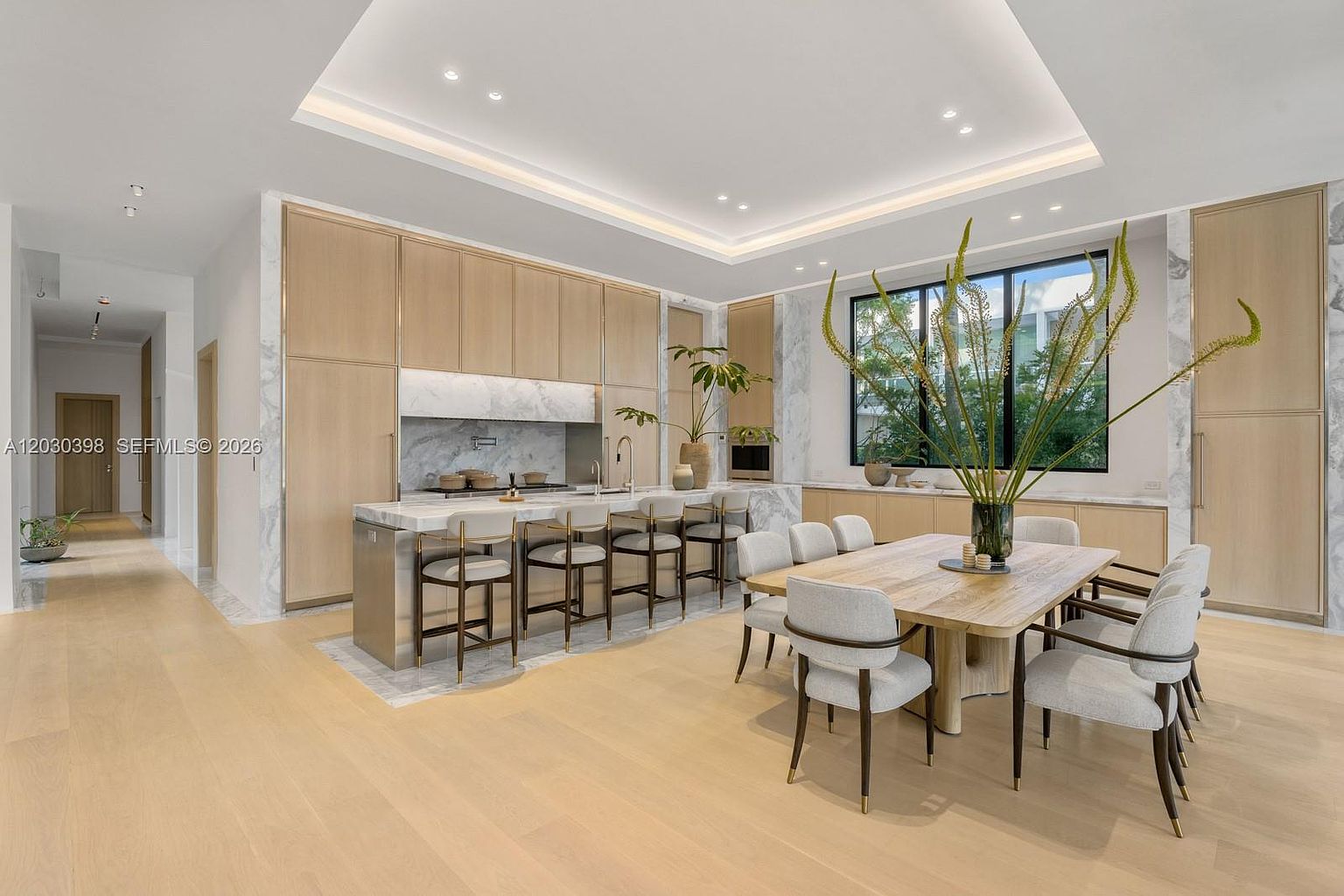 This bright, open-concept kitchen and dining area features light wood cabinetry and a large central island with stone countertops and bar seating. A dining table with upholstered chairs sits adjacent to the island, positioned near large windows that allow for ample natural light. The space is defined by a clean, modern aesthetic with neutral tones and recessed ceiling lighting, creating an inviting and airy atmosphere.