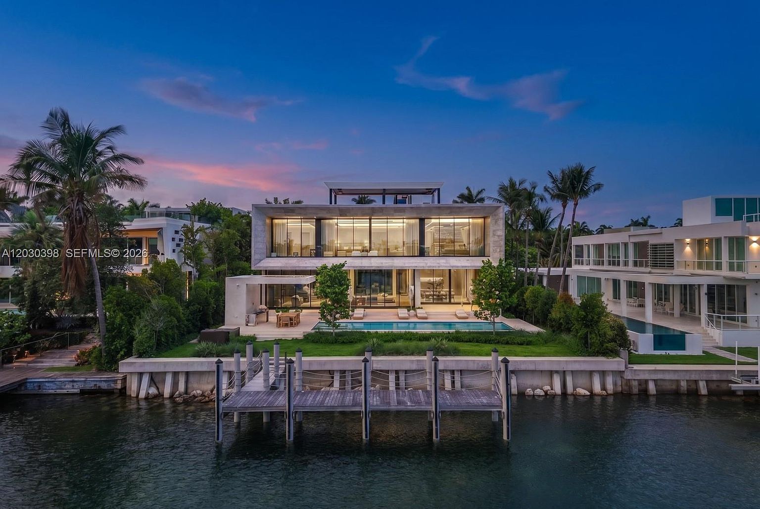 This stunning modern waterfront estate is captured from an elevated perspective, showcasing its sleek, geometric architecture and expansive glass walls. The property features a private dock extending into the calm water, a lush green lawn, and a rectangular swimming pool positioned directly in front of the residence. The twilight sky provides a dramatic backdrop, highlighting the warm interior lighting that glows through the large windows of the two-story structure.