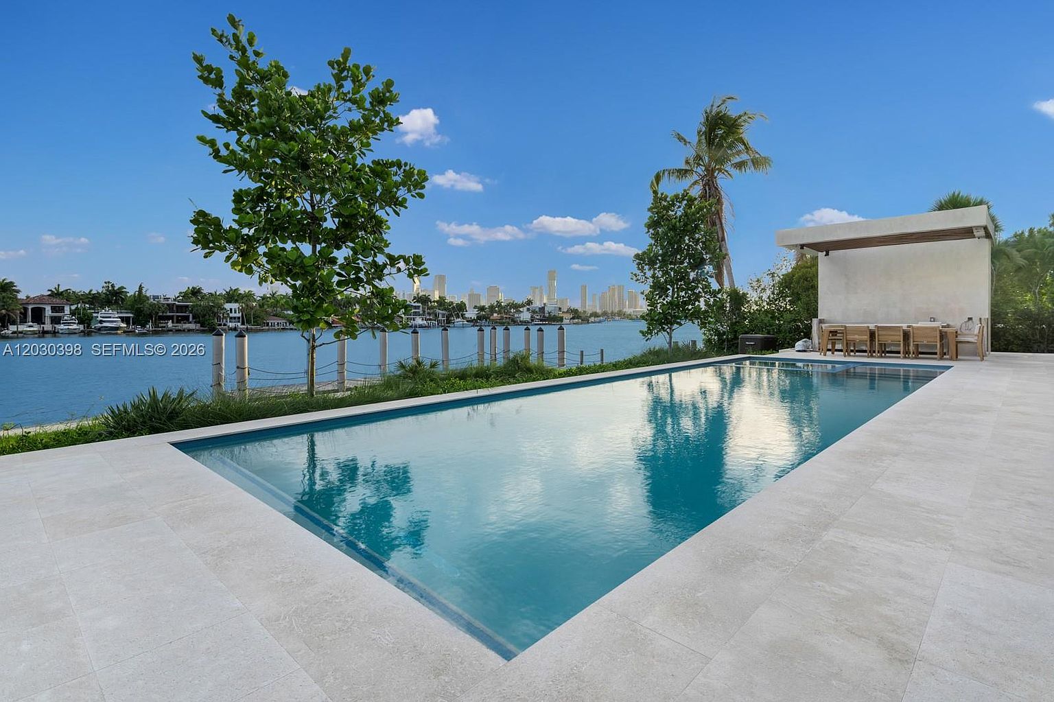 This image captures a serene outdoor living space featuring a rectangular swimming pool with clear blue water, set against a backdrop of a calm waterway and a distant city skyline. To the right, a covered patio area provides a shaded dining space with a long table and chairs, while lush green trees and a manicured lawn frame the water's edge. The perspective is from the pool deck, looking outward toward the horizon, emphasizing the property's waterfront location and tranquil atmosphere.