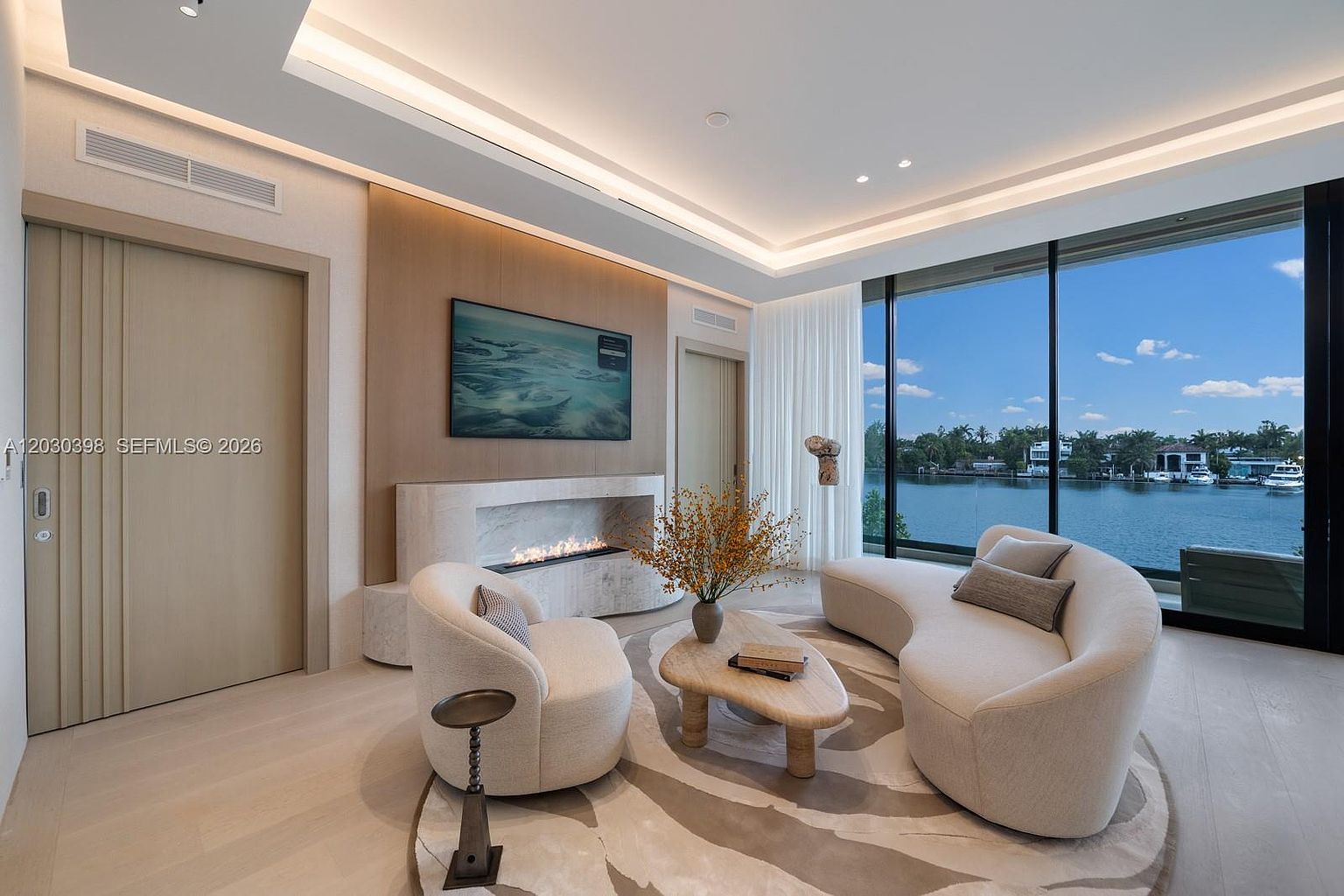 This sophisticated living room features a curved sofa and a matching armchair arranged around a unique, organic-shaped coffee table on a patterned area rug. A linear fireplace is set into a stone-clad wall, positioned beneath a wall-mounted television, while large windows provide an expansive view of the waterfront. The room is finished with light wood flooring and recessed ceiling lighting, creating a serene and luxurious atmosphere.