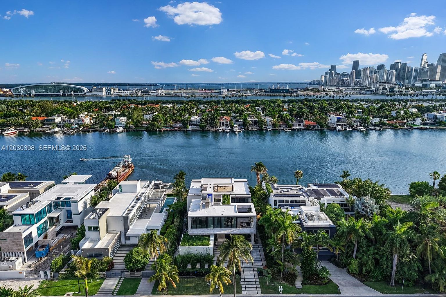 This high-angle aerial view captures a cluster of modern, multi-level waterfront residences along a canal, showcasing their contemporary architectural design and flat rooflines. The scene highlights the proximity to the water, with private docks and boats visible, while the background features a panoramic view of a distant city skyline under a bright, clear sky. The composition emphasizes the luxury lifestyle and prime location of these properties.