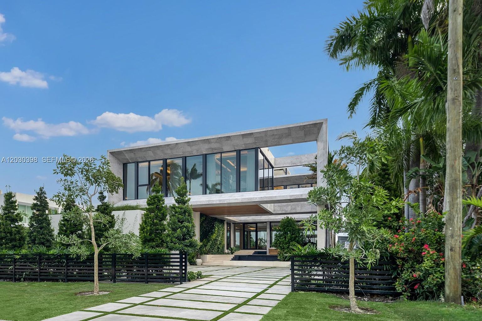 This striking modern residence features a bold, geometric architectural design with a prominent concrete frame and extensive glass walls. A paved walkway leads through a manicured lawn toward the recessed entryway, which is framed by lush landscaping and mature palm trees. The clean lines and open-concept aesthetic create a sophisticated, high-end impression of a contemporary luxury home.