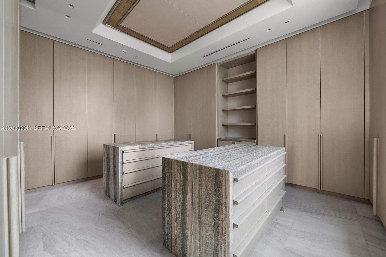 This spacious walk-in closet features light wood cabinetry and two central islands topped with patterned stone. The room is designed with a clean, minimalist aesthetic, incorporating open shelving and integrated lighting within a recessed ceiling detail. The perspective is centered, showcasing the symmetrical layout and high-end finishes of the storage space.