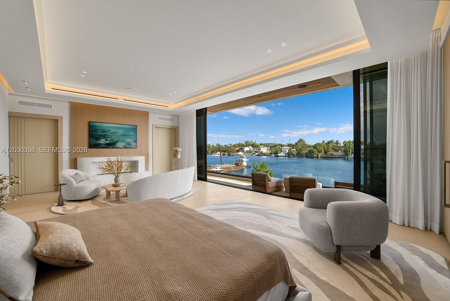 This luxurious primary bedroom features a contemporary design with a neutral color palette and expansive floor-to-ceiling glass walls that provide panoramic views of the waterfront. The room includes a cozy seating area with curved furniture, a television mounted above a sleek, minimalist fireplace, and a plush area rug that anchors the space. The perspective captures the seamless transition between the sophisticated interior and the serene outdoor canal setting.